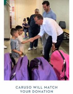 Man in white shirt shakes hands with a young boy near a row of backpacks during a donation event.