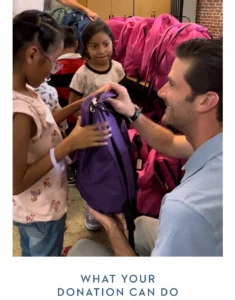 Man hands a purple backpack to a girl during a donation event, with pink backpacks arranged in the background.